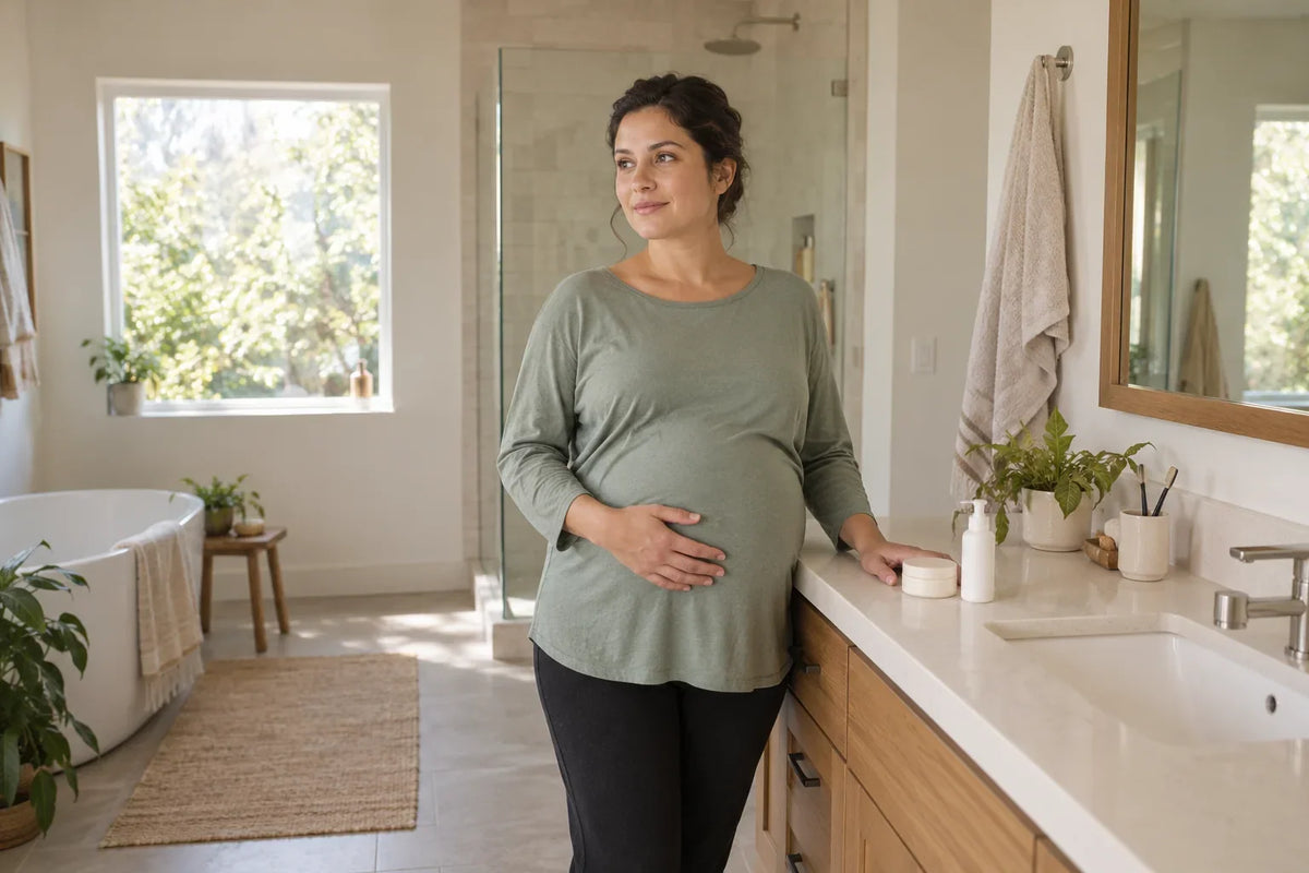 A pregnant woman in a green top stands beside a bathroom counter with a closed jar and a white pump bottle.